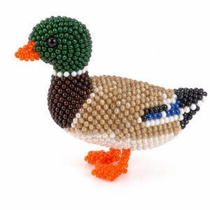 May include: A mallard duck figurine crafted from small, round beads. The head is dark green with a black eye and orange beak, the neck has a white ring, the chest is dark brown, and the body features tan, white, blue, and black bead patterns.