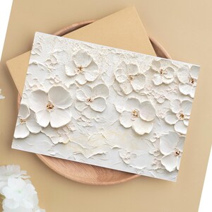 May include: A white greeting card with a textured floral design featuring raised white flowers with gold centers. The card is placed on a wooden tray with a brown envelope.