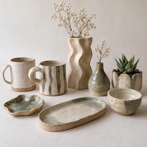 May include: A collection of handmade ceramic items including two mugs, a wavy vase, a small bottle vase, a scalloped planter with a succulent, a small dish, an oval tray, and a bowl, all featuring speckled cream and sage green glazed finishes.