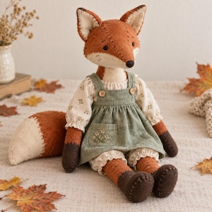 Può includere: Una bambola volpe di peluche con pelliccia color ruggine, viso color crema e una grande coda folta. La volpe indossa una camicetta color crema con un piccolo motivo floreale sotto un vestito con pettorina verde salvia con tasca ricamata.