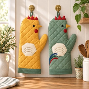 Puede incluir: Dos manoplas de cocina acolchadas con forma de gallina colgadas de ganchos dorados. Una manopla es amarilla con cresta roja y ala blanca, mientras que la otra es verde salvia con cresta roja, ala blanca y plumas de cola azules y rojas.