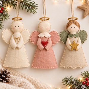 May include: Three handcrafted felt angel ornaments hanging by twine. The left angel is cream with a gold halo and bow. The centre angel is dusty pink with a red heart. The right angel has brown yarn hair, green wings, and a gold star.