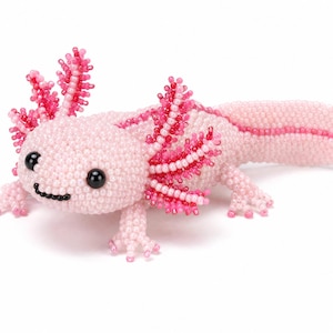 Könnte beinhalten: Eine perlenbesetzte Axolotl-Figur aus kleinen, hellrosa Perlen. Das Wesen hat markante, leuchtend dunkelrosa Außenkiemen, zwei große schwarze Perlenaugen und einen kleinen, schwarz gestickten Mund, mit einem langen Schwanz nach rechts.