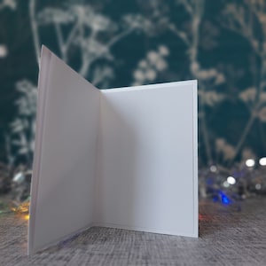 May include: A blank, white, rectangular greeting card standing upright in an open position. The card features a clean, smooth surface on the interior panels, providing a neutral space for writing or printing messages on both sides of the fold.