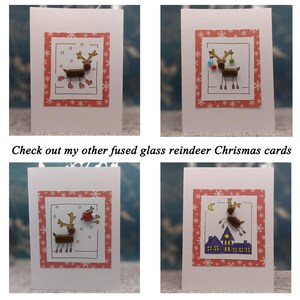 May include: Four white greeting cards featuring fused glass reindeer designs. Each card has a red border with white snowflakes. The reindeer are depicted in various scenes including ice skating, holding ornaments, with a bird, and flying over houses.