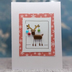 May include: A white greeting card featuring a central illustration of a brown reindeer with tan antlers. The reindeer is decorated with small, colorful hanging beads in red, light blue, and green. The design is framed by a red border with white snowflakes.