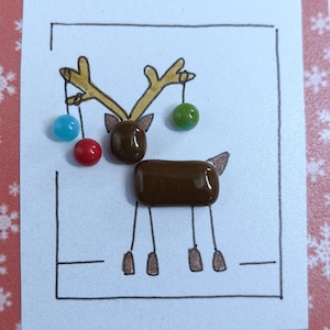 May include: A white card featuring a hand-drawn reindeer with brown glass cabochon pieces for the head and body. The reindeer has ink-drawn antlers decorated with three small, round, colorful glass beads in light blue, red, and green.
