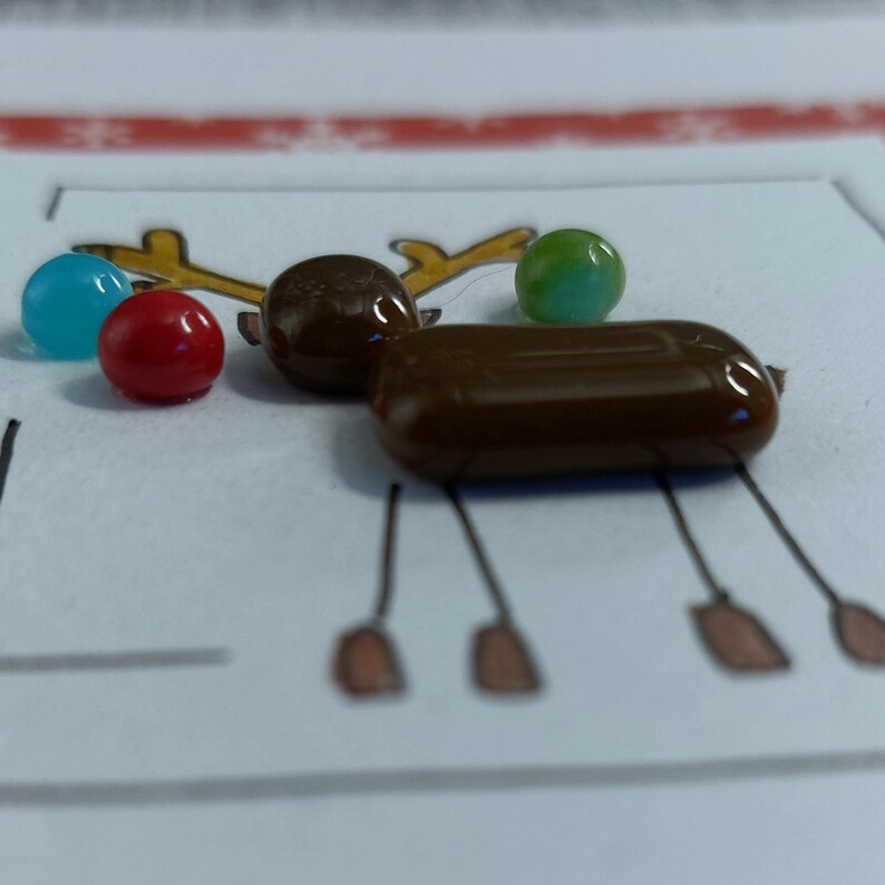 May include: A collection of small, smooth, glossy candy pieces arranged on a white paper surface featuring a hand-drawn illustration of a reindeer. The candies include a blue sphere, a red sphere, a green sphere, a brown sphere, and a brown oval.