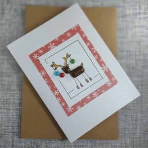 May include: A white greeting card featuring a central illustration of a reindeer with brown glass accents for the head and body, gold ink antlers, and small colorful glass beads. The card is framed by a red border with white snowflake patterns.