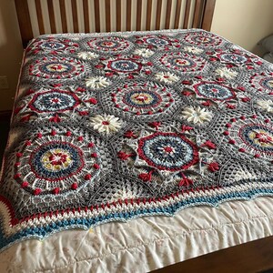 K&ouml;nnte beinhalten: Eine geh&auml;kelte Decke mit grauem Hintergrund und bunten kreisf&ouml;rmigen Mustern in Rot, Wei&szlig;, Blau und Gelb. Die Decke liegt auf einem Bett mit einem wei&szlig;en Bettbezug.