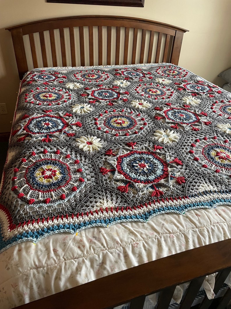 K&ouml;nnte beinhalten: Eine geh&auml;kelte Decke mit grauem Hintergrund und bunten kreisf&ouml;rmigen Mustern in Rot, Wei&szlig;, Blau und Gelb. Die Decke liegt auf einem Bett mit einem wei&szlig;en Bettbezug.