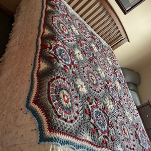 K&ouml;nnte beinhalten: Eine graue, wei&szlig;e, rote und blaue geh&auml;kelte Decke mit geometrischem Muster. Die Decke liegt auf einem Bett mit einer wei&szlig;en Bettw&auml;sche mit Blumenmuster.