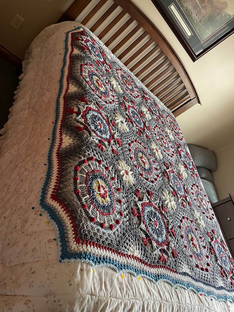 K&ouml;nnte beinhalten: Eine graue, wei&szlig;e, rote und blaue geh&auml;kelte Decke mit geometrischem Muster. Die Decke liegt auf einem Bett mit einer wei&szlig;en Bettw&auml;sche mit Blumenmuster.