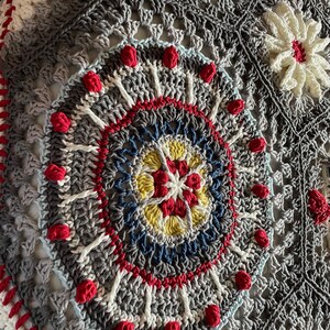 K&ouml;nnte beinhalten: Nahaufnahme einer geh&auml;kelten Decke mit einem kreisf&ouml;rmigen Muster in Graut&ouml;nen, Wei&szlig;, Rot, Blau und Gelb. Die Mitte des Kreises zeigt ein kleineres, mehrfarbiges Sternmuster.