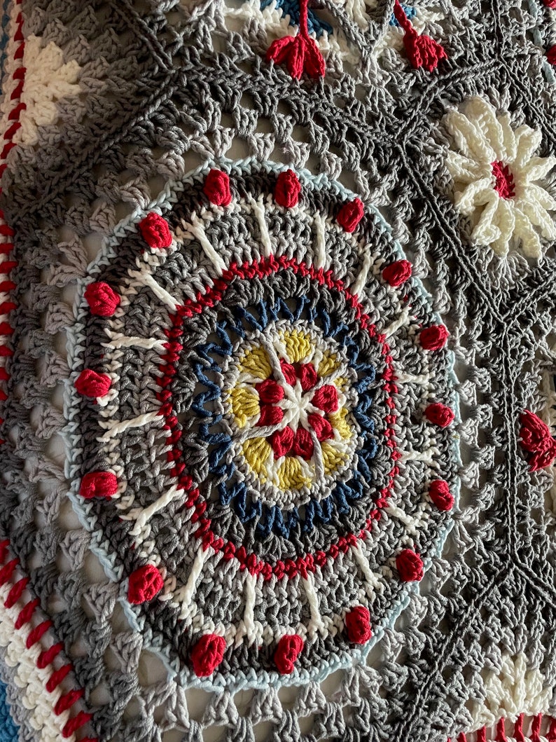 K&ouml;nnte beinhalten: Nahaufnahme einer geh&auml;kelten Decke mit einem kreisf&ouml;rmigen Muster in Graut&ouml;nen, Wei&szlig;, Rot, Blau und Gelb. Die Mitte des Kreises zeigt ein kleineres, mehrfarbiges Sternmuster.