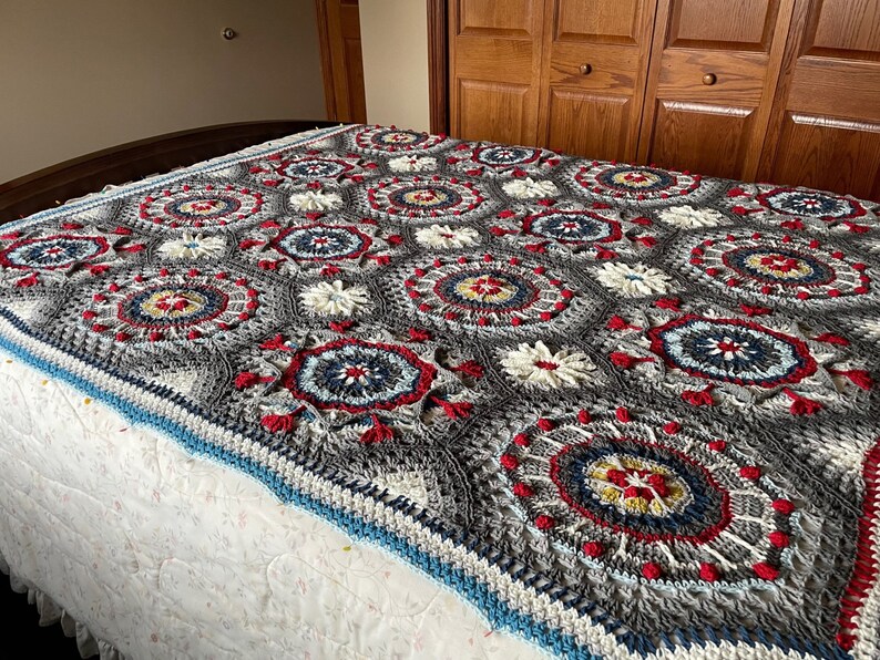 K&ouml;nnte beinhalten: Eine geh&auml;kelte Decke mit grauem Hintergrund und bunten kreisf&ouml;rmigen Mustern in Rot, Wei&szlig;, Blau und Gelb.