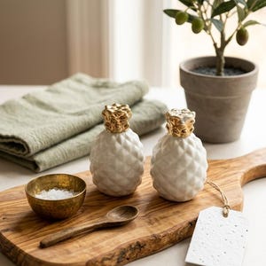 Puede incluir: Dos saleros de cerámica blanca con forma de piña y tapas de oro metálico, un pequeño cuenco de latón con sal, una cuchara de madera y una etiqueta de papel blanco con cuerda de yute, dispuestos sobre una tabla de cortar de madera natural.