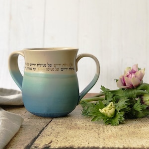 Puede incluir: Una taza de cerámica azul y blanca con dos asas y escritura en hebreo. La taza está sobre una superficie de madera con un ramo de flores rosas y blancas.