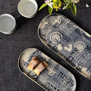 Könnte beinhalten: Zwei ovale Servierplatten aus Keramik mit einem schwarz-weißen Design mit hebräischem Text und abstrakten Mustern. Die Platten befinden sich auf einer dunkelgrauen Oberfläche mit einem kleinen Strauß weißer und gelber Blumen in einem Topf.