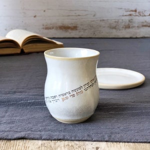 May include: White ceramic Kiddush cup with Hebrew text in gold and brown. The cup is sitting on a gray tablecloth with a white saucer behind it.