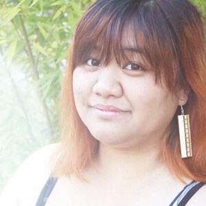 May include: A woman with long, reddish-brown hair wearing a black tank top and long, rectangular earrings with a gold and white pattern.