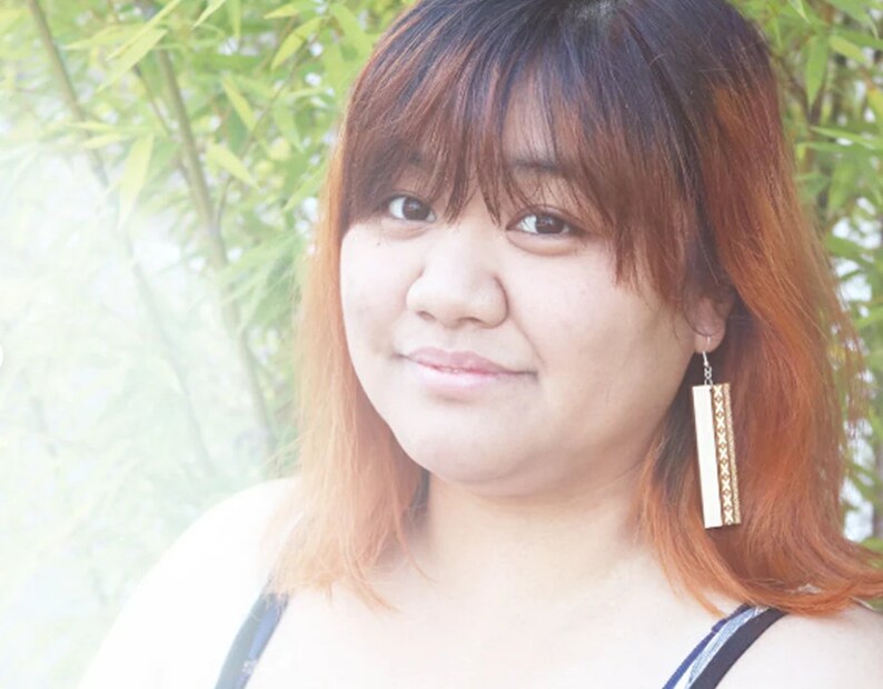 May include: A woman with long, reddish-brown hair wearing a black tank top and long, rectangular earrings with a gold and white pattern.