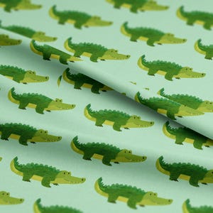 May include: A light green fabric with a repeating pattern of cartoon alligators. The alligators are green with yellow bellies and are arranged in a diagonal pattern across the fabric. The fabric has a soft, matte finish.