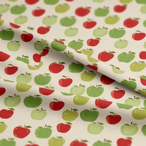 May include: Cream-coloured fabric featuring a repeating pattern of red, green, and yellow apples. The apples are whole and sliced, with stems and leaves. The design has a whimsical, hand-drawn aesthetic.