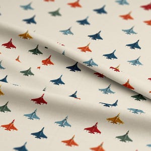 Puede incluir: Tela de color crema con un patrón repetido de aviones de combate coloridos. Los aviones son rojos, naranjas, amarillos, verdes y azules. La tela tiene una textura sutil y está doblada en diagonal.