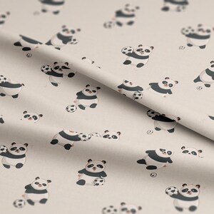 May include: Cream-coloured fabric featuring a repeating pattern of cartoon pandas playing football. The pandas are black and white with small footballs. The fabric is folded, showing the pattern's continuity.