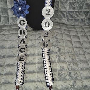 May include: A graduation lei in white, navy blue, and silver. The lei is braided with white and navy blue ribbons. It features the name "GRACE" and the year "2026" in silver letters and numbers. A navy blue flower with a rhinestone center is also included.