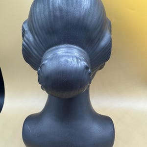 Puede incluir: Una cabeza de maniqu&iacute; gris oscuro con un peinado cl&aacute;sico, vista desde atr&aacute;s. La cabeza y el cuello son lisos, con el pelo peinado en un mo&ntilde;o y ondas. El maniqu&iacute; est&aacute; dise&ntilde;ado para exhibir pelucas, sombreros o joyas.
