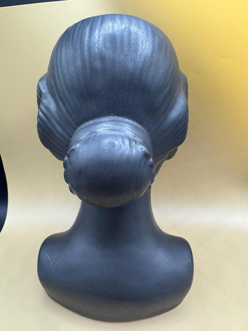 Puede incluir: Una cabeza de maniqu&iacute; gris oscuro con un peinado cl&aacute;sico, vista desde atr&aacute;s. La cabeza y el cuello son lisos, con el pelo peinado en un mo&ntilde;o y ondas. El maniqu&iacute; est&aacute; dise&ntilde;ado para exhibir pelucas, sombreros o joyas.