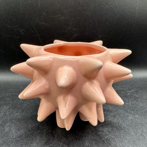 May include: A light pink ceramic planter featuring a spherical body covered in numerous protruding, cone-shaped spikes. The vessel has a wide, circular opening at the top and a smooth, glossy finish across its entire exterior surface.