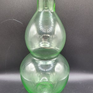 May include: A green glass vase shaped like a gourd with two rounded sections. The vase is clear and has a slight frosted appearance.