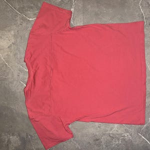 May include: A coral-colored t-shirt with a crew neck and short sleeves. The shirt is made of a soft material and has a pocket on the left chest. The shirt is laid flat on a gray surface.