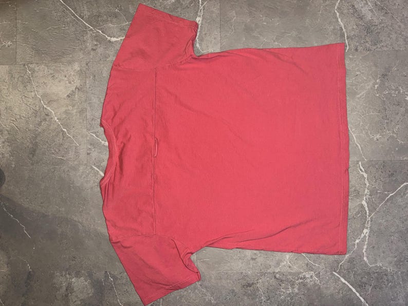 May include: A coral-colored t-shirt with a crew neck and short sleeves. The shirt is made of a soft material and has a pocket on the left chest. The shirt is laid flat on a gray surface.