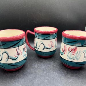 May include: Three ceramic mugs with a cream-colored interior, featuring a teal and white striped design with pink floral accents. The rims and handles are a deep red. The mugs are arranged on a dark surface.