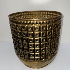 May include: A cylindrical brass planter featuring a repeating geometric pattern of raised squares with central indentations. The metal surface has a warm, aged patina with a smooth, plain band at the base and a decorative scalloped rim.