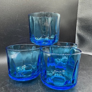 May include: Four blue drinking glasses with a textured, ridged design. The glasses are a vibrant shade of blue and appear to be made of glass. The glasses are round and have a thick base. The glasses are empty.