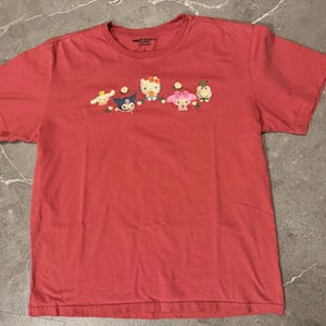 May include: A coral-colored t-shirt featuring an embroidered design of several cartoon characters. The characters include a white cat with a red bow, a blue character, and other figures with floral accents. The shirt has short sleeves and a crew neck.