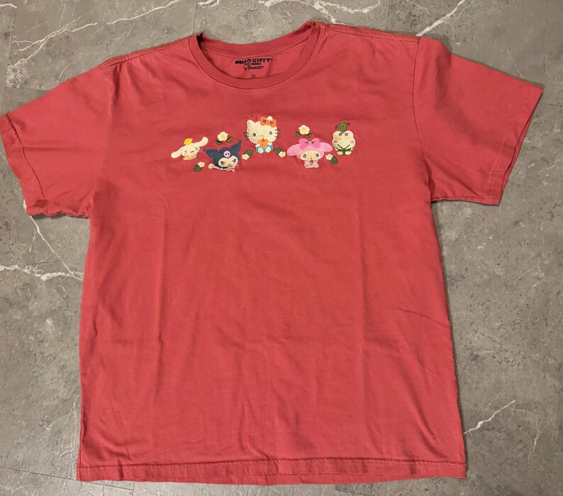 May include: A coral-colored t-shirt featuring an embroidered design of several cartoon characters. The characters include a white cat with a red bow, a blue character, and other figures with floral accents. The shirt has short sleeves and a crew neck.