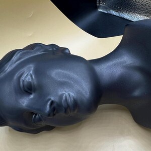 Puede incluir: Cabeza de maniqu&iacute; negra mate que representa un busto femenino cl&aacute;sico. La cabeza est&aacute; inclinada hacia la izquierda, con los ojos cerrados y rasgos detallados. El busto es una forma s&oacute;lida y lisa.
