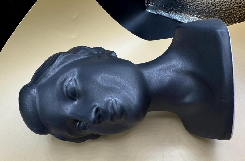 Puede incluir: Cabeza de maniqu&iacute; negra mate que representa un busto femenino cl&aacute;sico. La cabeza est&aacute; inclinada hacia la izquierda, con los ojos cerrados y rasgos detallados. El busto es una forma s&oacute;lida y lisa.