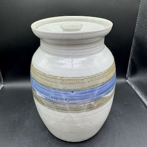 Może przedstawiać: Ręcznie robiony ceramiczny słoik z pokrywką. Słoik jest głównie biały z poziomymi pasami w kolorze niebieskim, brązowym i szarym. Pokrywka ma pasujący, kremowy kolor. Słoik ma zaokrąglony kształt i szeroki otwór.