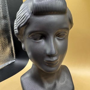 Puede incluir: Busto negro mate de una persona con cabello y rasgos faciales estilizados. La escultura tiene un acabado liso y est&aacute; sobre un fondo dorado. El busto es un objeto decorativo.