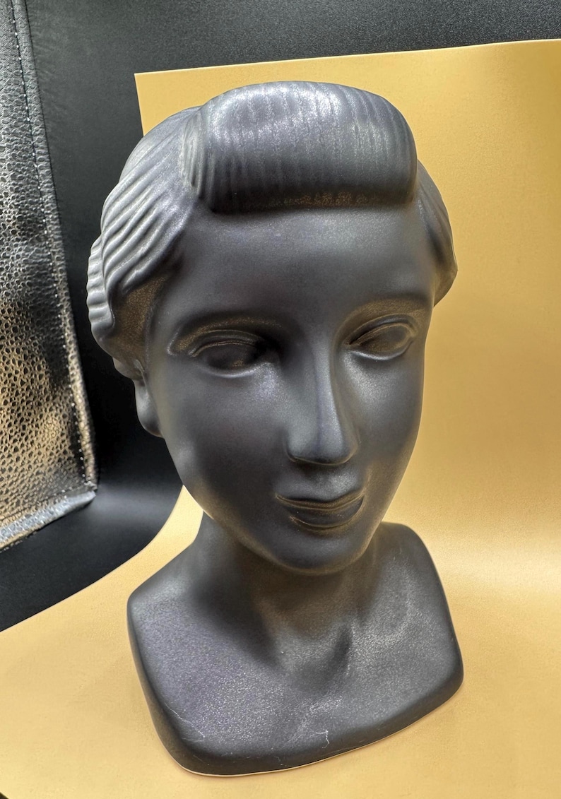 Puede incluir: Busto negro mate de una persona con cabello y rasgos faciales estilizados. La escultura tiene un acabado liso y est&aacute; sobre un fondo dorado. El busto es un objeto decorativo.