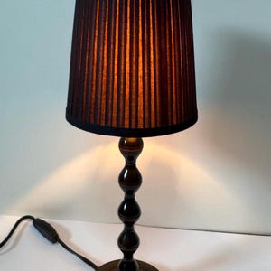 Op de afbeelding: Een tafellamp met een donkerbruine, verticaal geplooide cilindrische kap en een slanke, donkerbruine voet met vier gestapelde, ronde, bolvormige segmenten. De lamp is voorzien van een zwart snoer met een schakelaar.