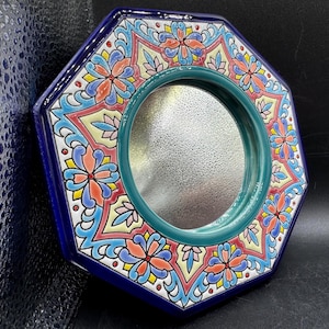 May include: An octagonal wall mirror featuring a deep blue outer frame and a central circular glass pane. The border is decorated with a colorful floral pattern in shades of coral, light blue, yellow, and cream, set against a dark blue background.