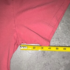 May include: Close-up of a coral-colored t-shirt sleeve with a yellow measuring tape. The tape shows a measurement of approximately 6 inches. The shirt's fabric is smooth and appears to be cotton.